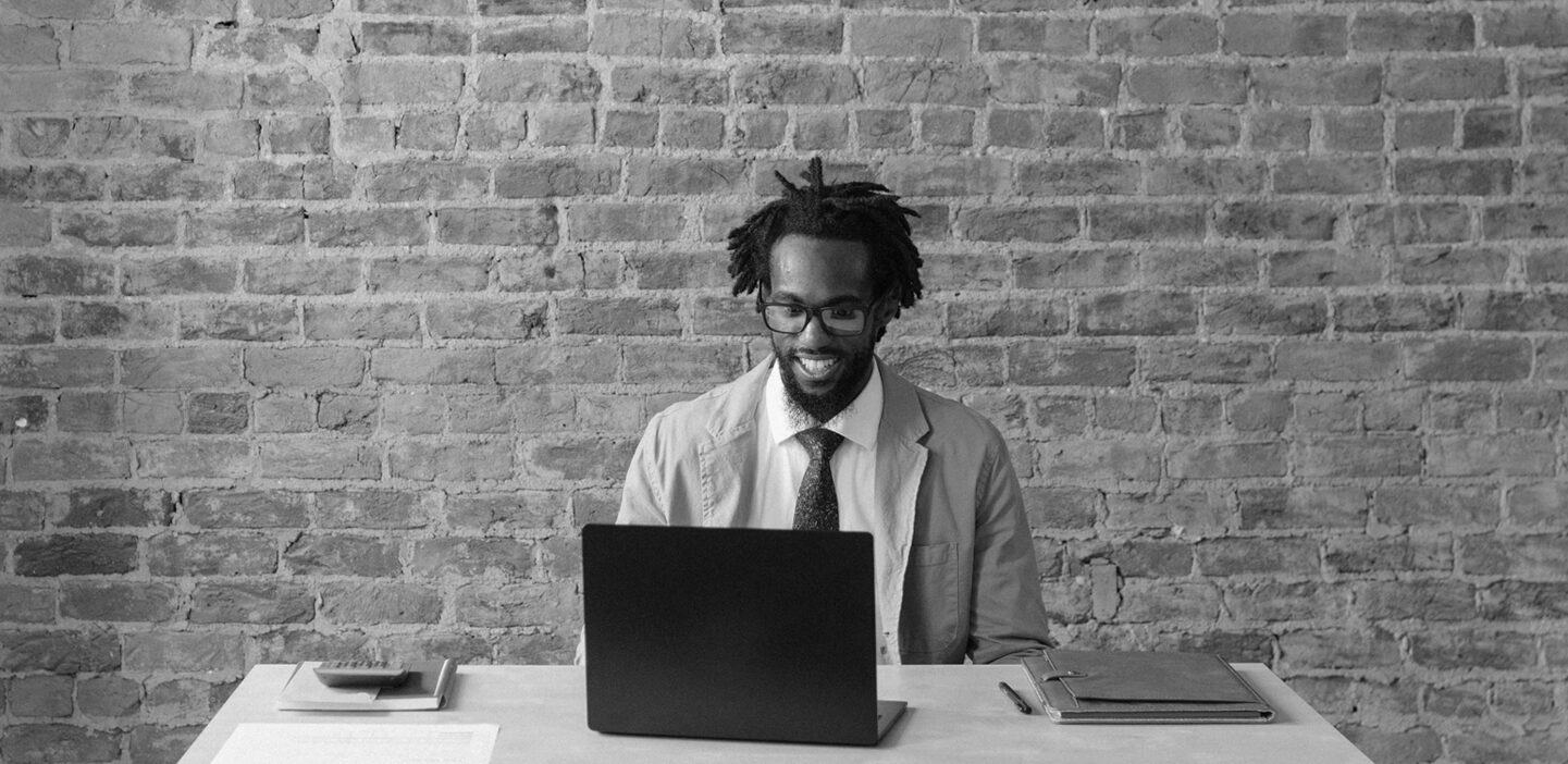 Man sitting at a desk in front of a brick wall, and working on a laptop