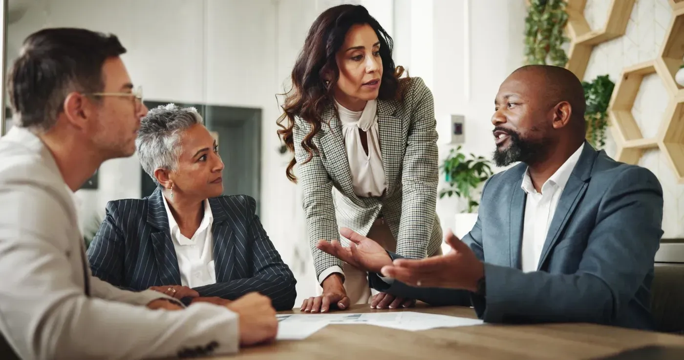 A female business leader standing at a conference table, actively leading a discussion with three diverse colleagues during a strategic meeting.