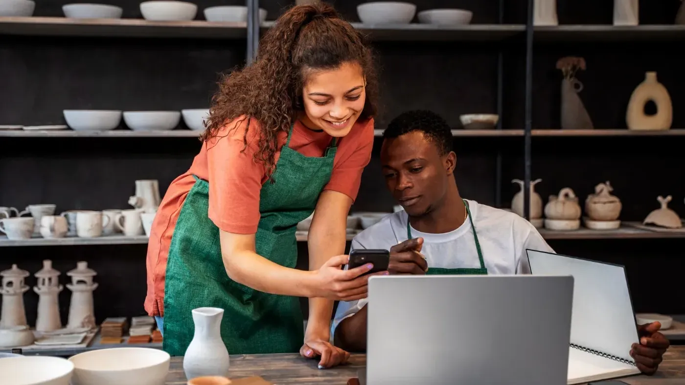 Two small business pottery professionals looking at a smart phone and laptop